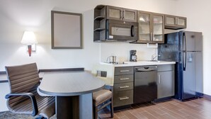 A compact kitchen with a microwave, refrigerator, and built-in cabinets. There is a dining table with chairs and a lamp on the wall. - Candlewood Suites Cut Off by IHG (Cut Off)