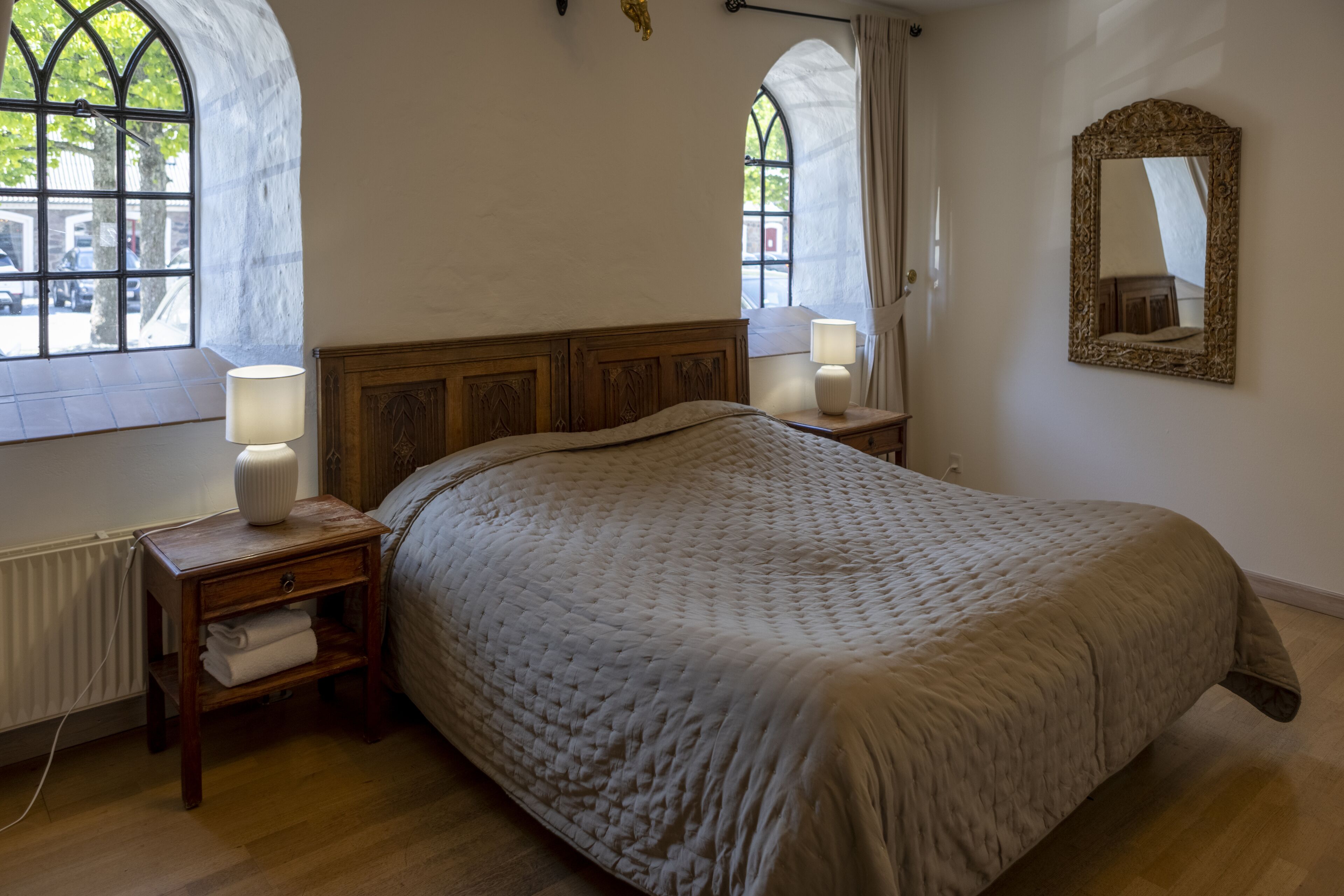Een slaapkamer met een groot bed, een houten hoofdbord, bedlampen, een nachtkastje en een raam met gordijnen.