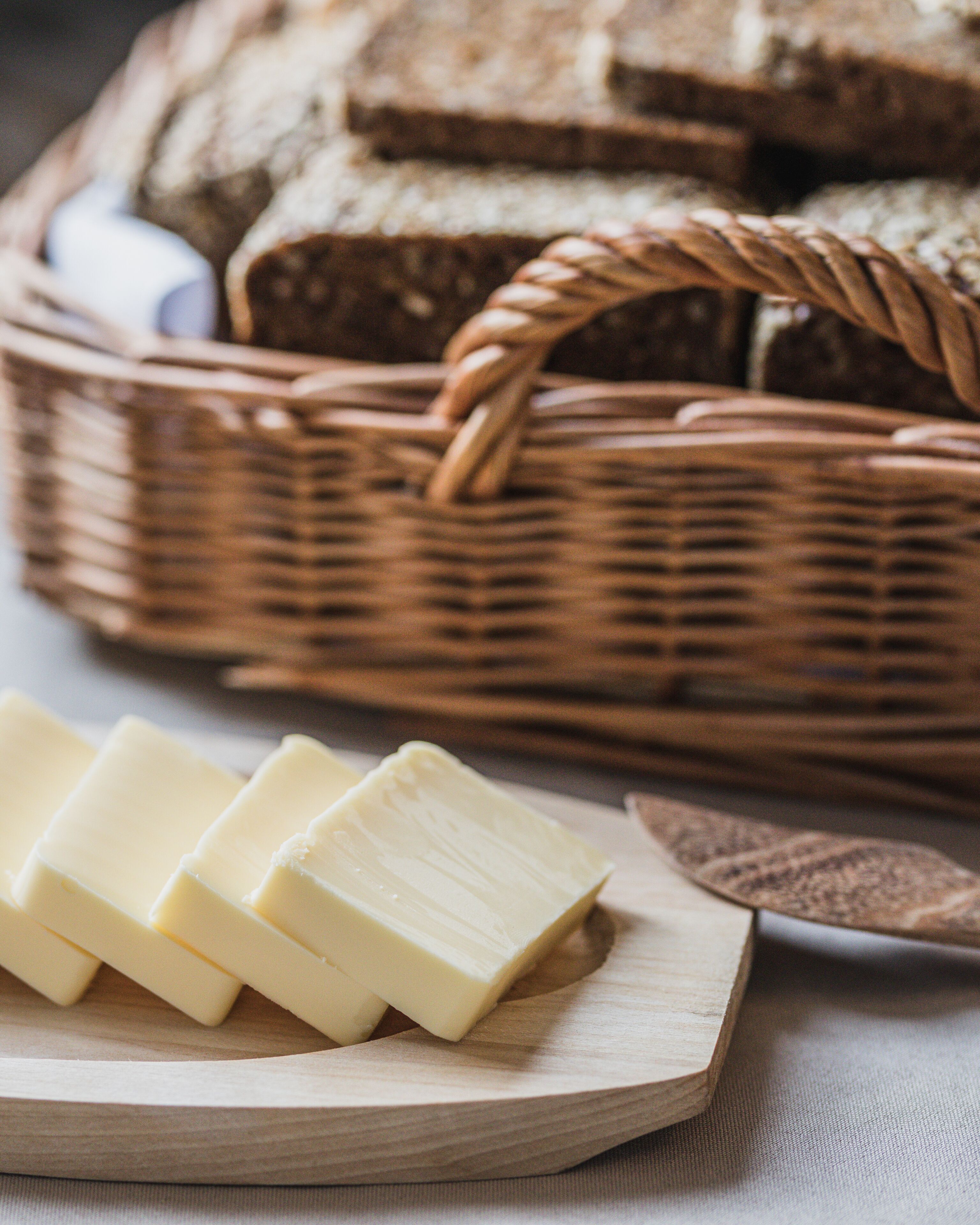 A wooden cutting board with several slices of butter and a wicker basket with bread in the background.