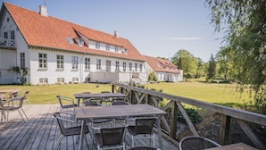 A wooden deck with tables and chairs, a white building with a red roof, and a grassy lawn. - Sonnerupgaard Gods (Hvalso)