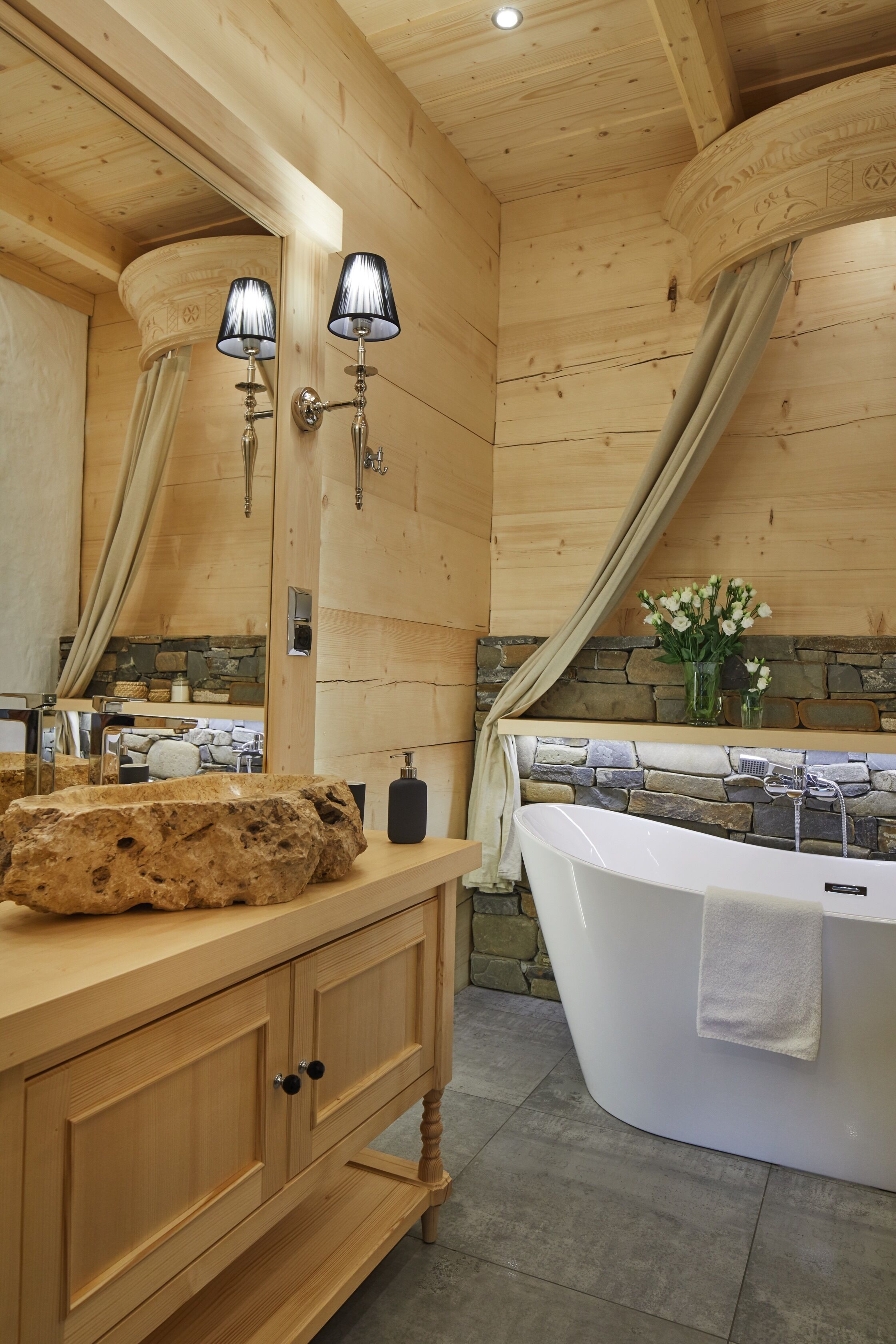 A wooden cabin bathroom with a stone wall, a bathtub, and a wooden vanity with a mirror and two wall-mounted lights.