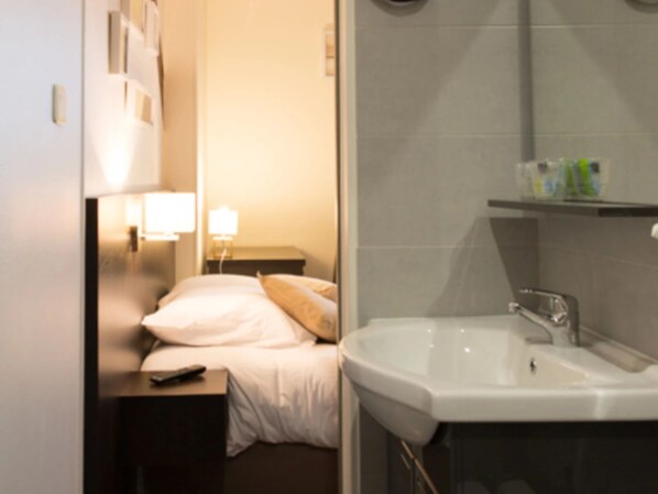 Habitación de hotel con cama, mesita de noche, lámpara y baño con lavamanos y espejo.