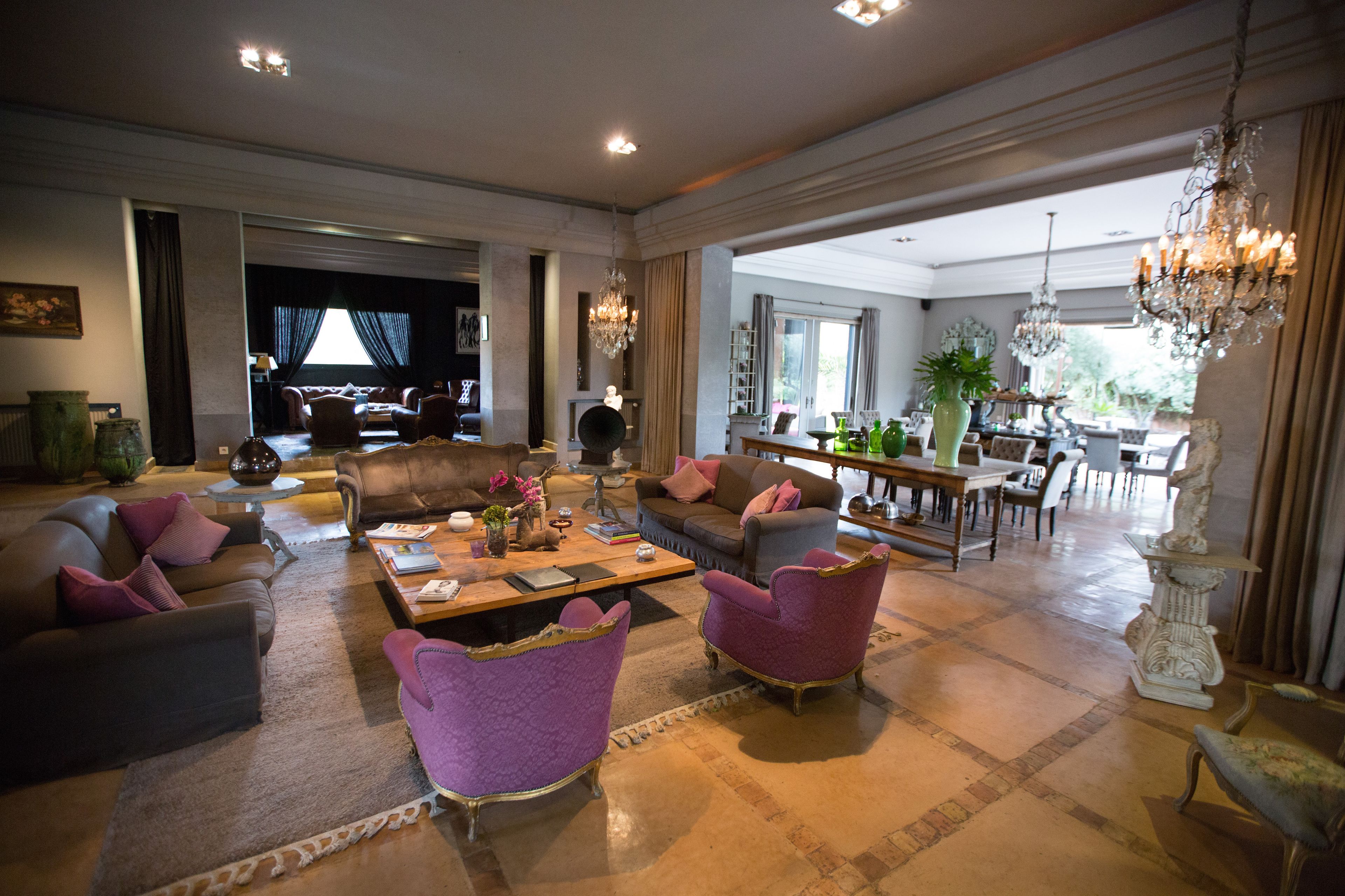 A spacious living room with a large coffee table, multiple sofas, and chairs. There are chandeliers and a dining area visible in the background.