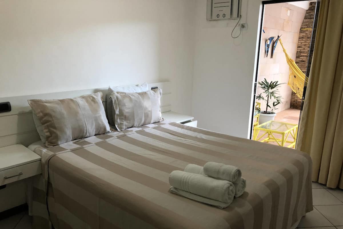 A hotel room with a large bed, two pillows, a bedside table, and a small table with a chair outside.