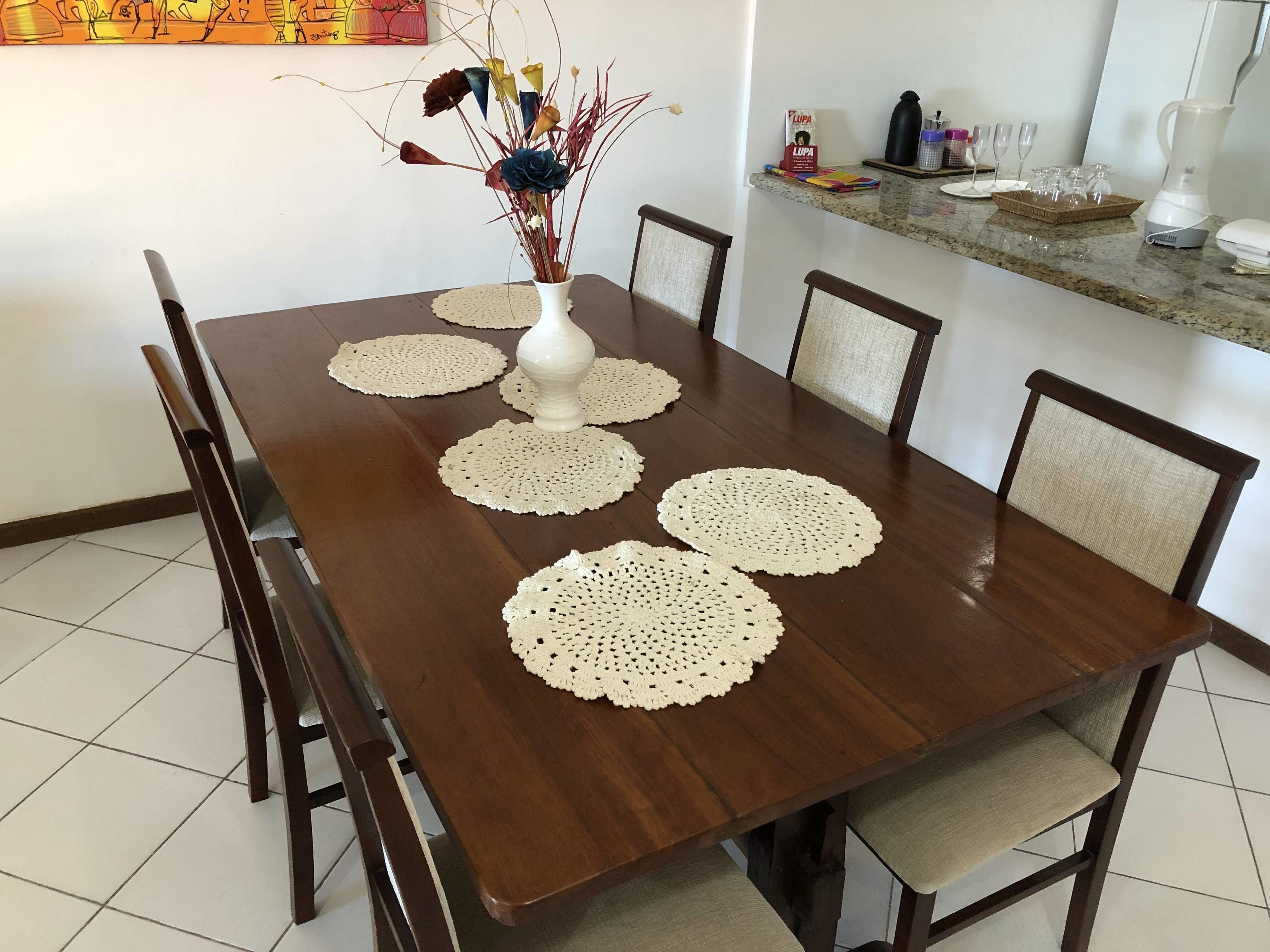 A dining table with six chairs, a vase with dried flowers, and a kitchen counter with various items.