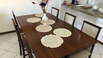 A dining table with six chairs, a vase with dried flowers, and a kitchen counter with various items.