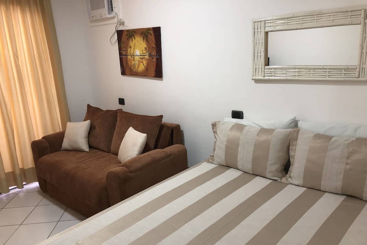 A room with a brown sofa, a striped bed, a wall-mounted picture, and a window with curtains.
