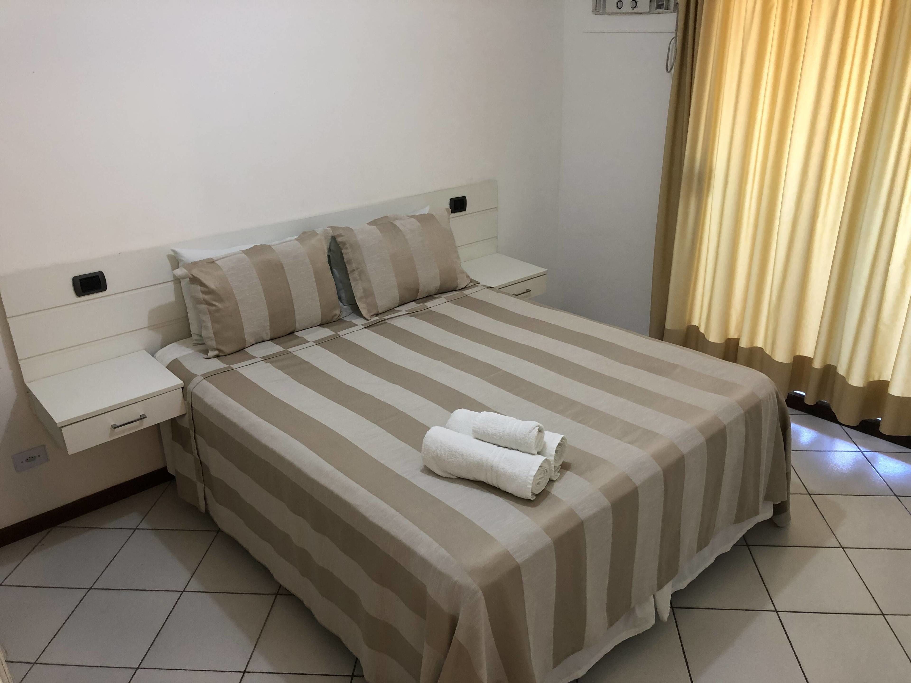 A neatly made bed with striped bedding, a bedside table with a drawer, and a rolled towel on the bed.