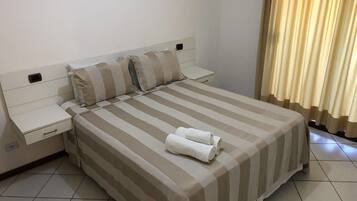 A neatly made bed with striped bedding, a bedside table with a drawer, and a rolled towel on the bed.