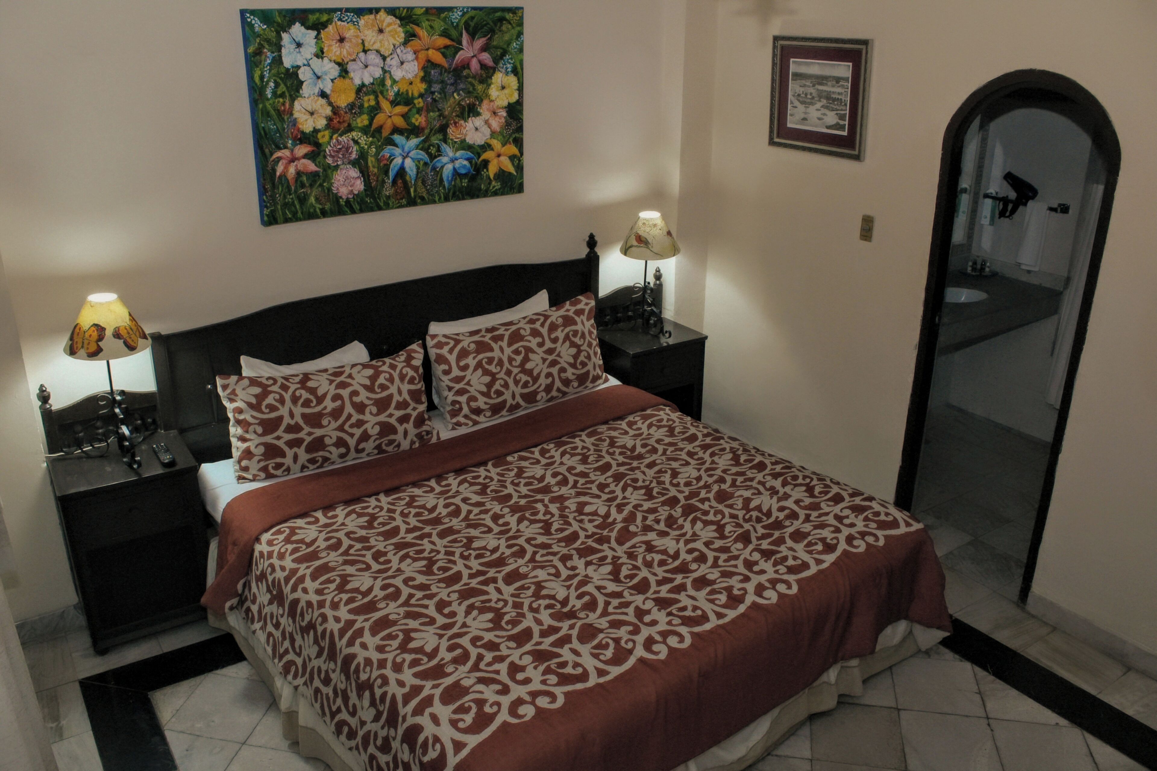 A bedroom with a bed, nightstands, a painting, a framed picture, and a mirror.
