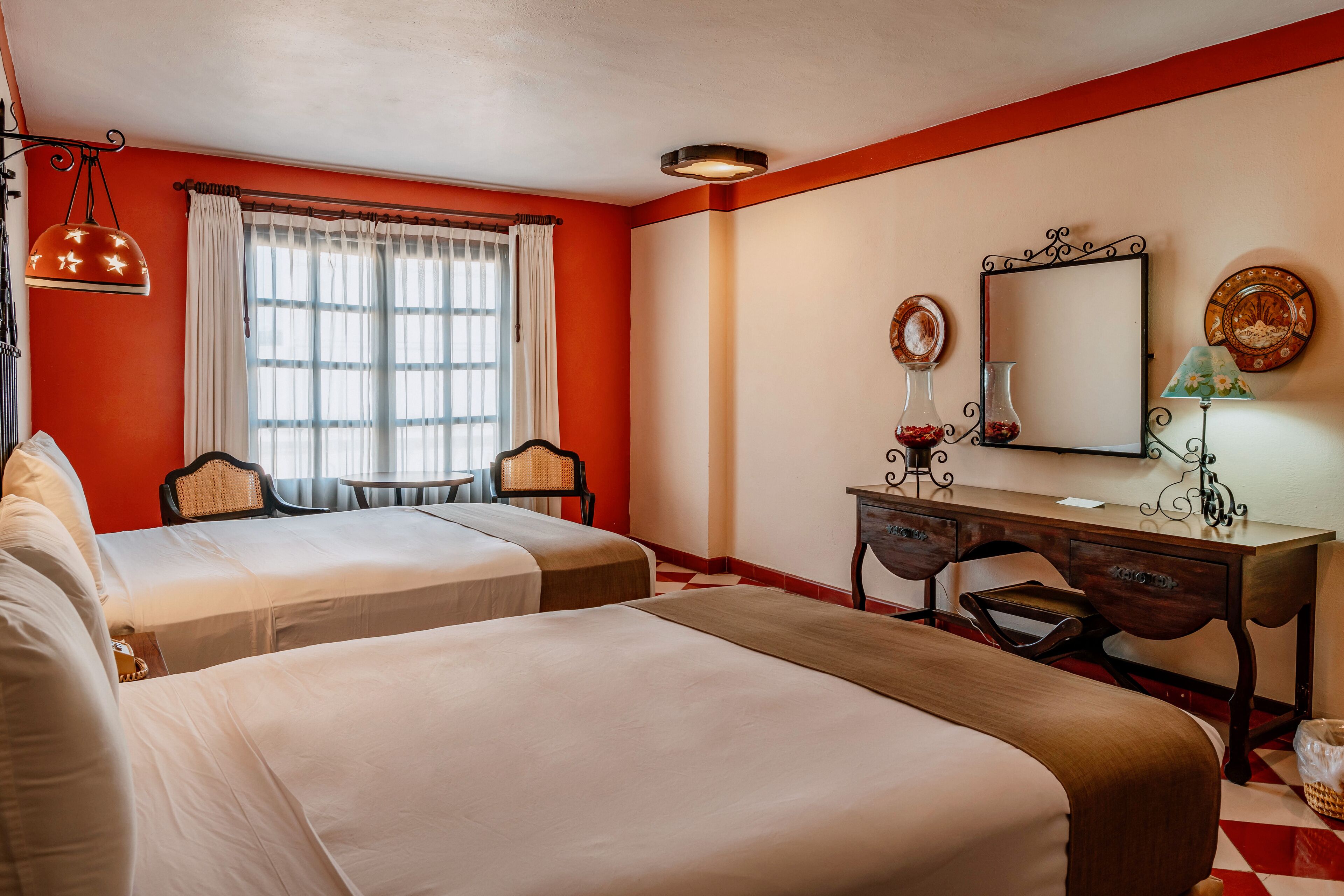 A hotel room with two beds, a desk, a chair, a lamp, and a window with curtains.