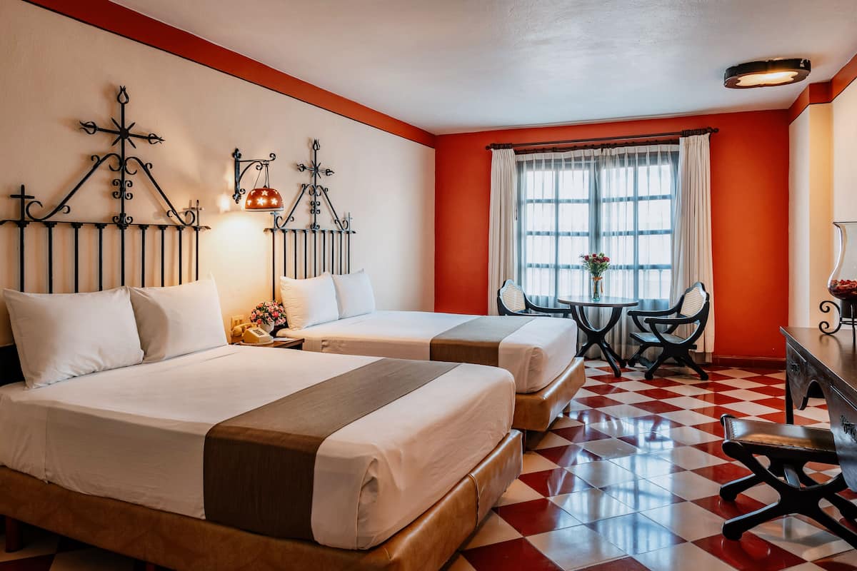 A hotel room with two beds, a dining table with chairs, a small table with a vase of flowers, and a window with curtains.