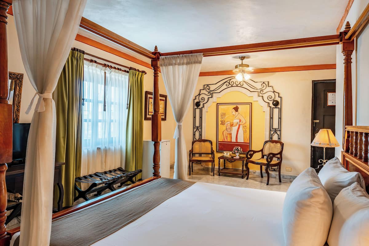 Habitación de hotel con una cama grande, un banco de madera, dos sillas, una mesa y un cuadro en la pared.