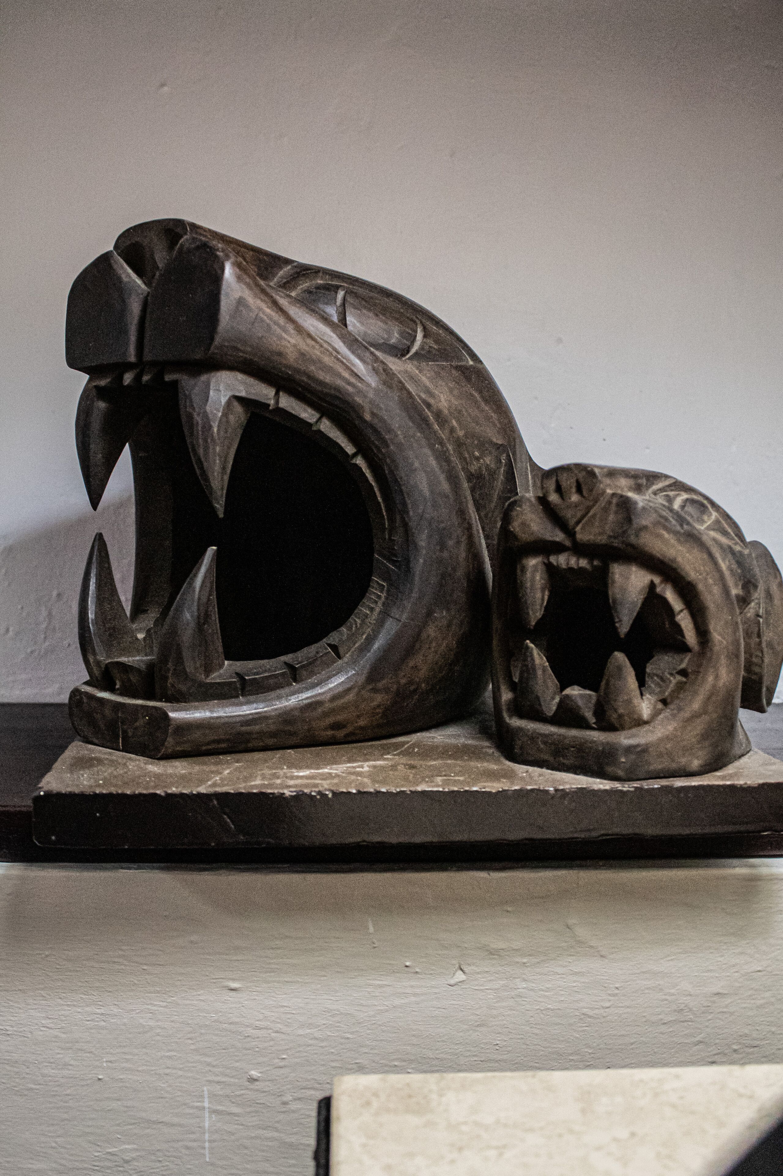 En skulptur af et brølende dyr, muligvis en løve, med skarpe tænder og kløer, placeret på en mørk overflade foran en enkel baggrund.