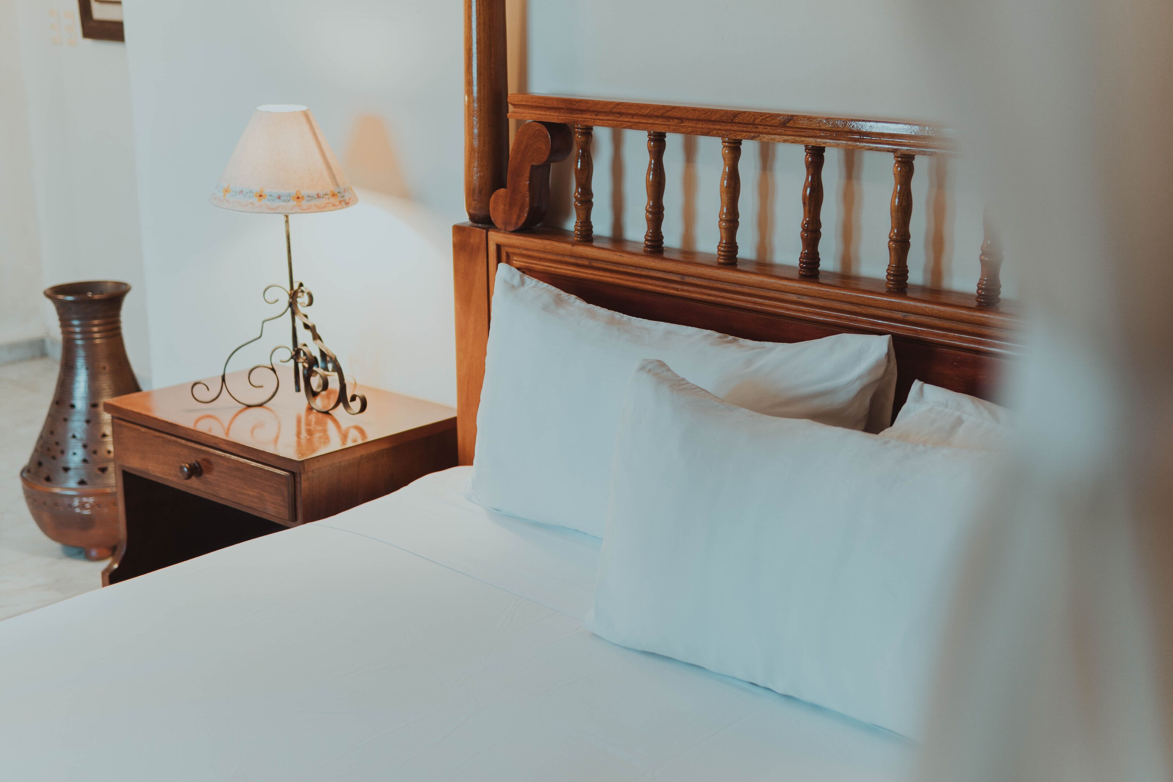 A neatly made bed with white linens, a wooden headboard, and a bedside table with a lamp and a vase.