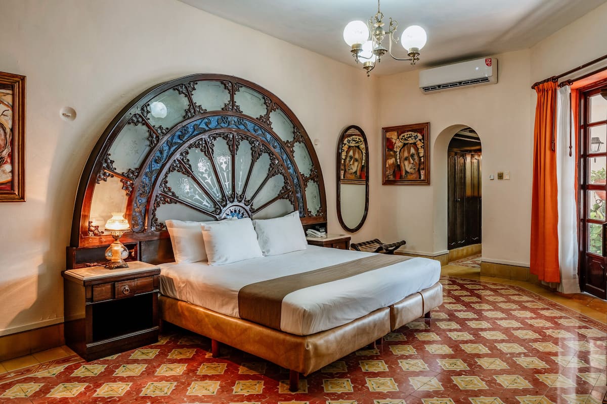A bedroom with a large bed, a nightstand, a lamp, a chandelier, and a large window with curtains.