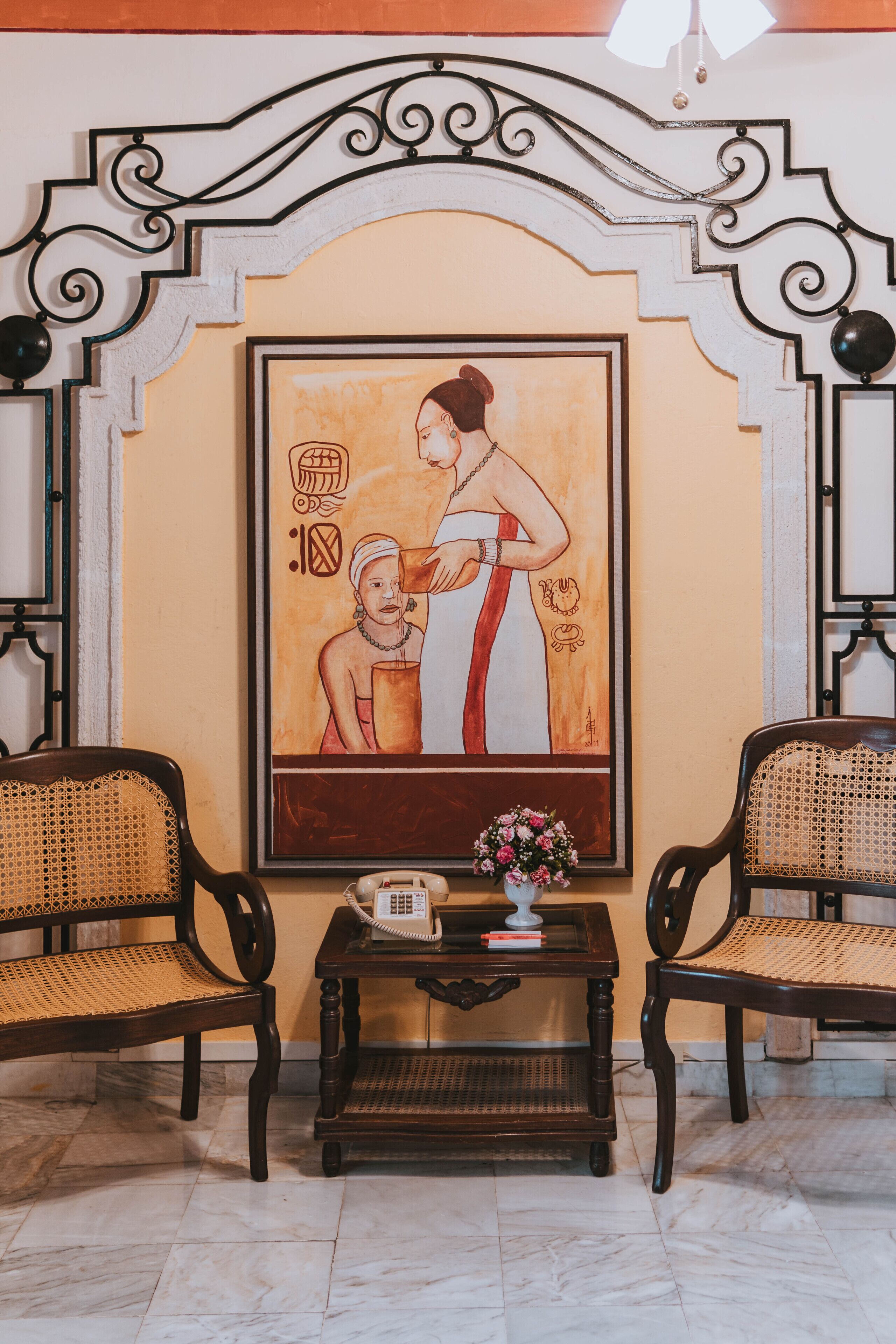 A room with two wicker chairs, a small table with a telephone and a vase of flowers, and a large framed painting on the wall.