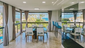 A spacious room with a large window overlooking a beach, a dining area with chairs and tables, and a kitchen area with a counter and chairs. - Villaggio Orizzonte (Salvador)