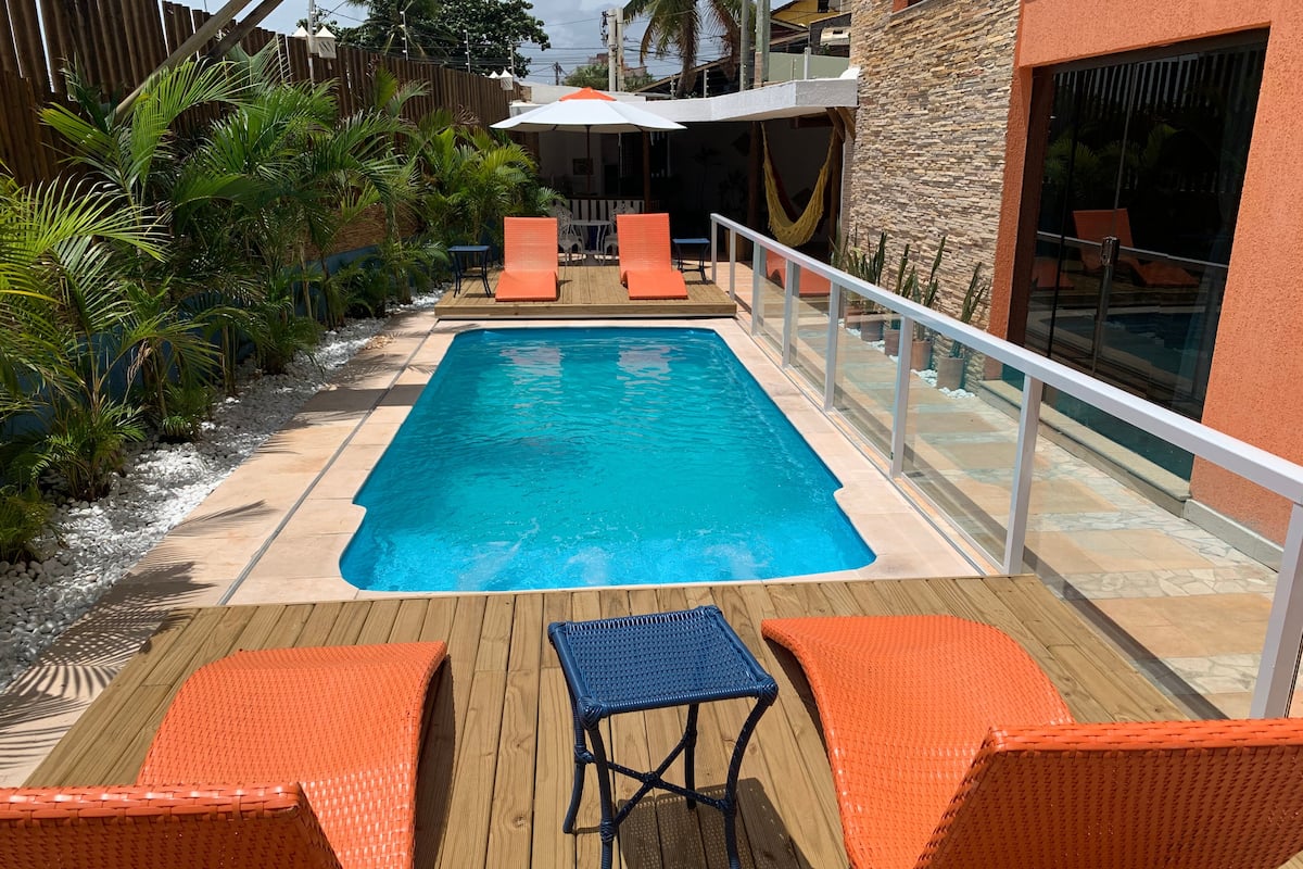 A swimming pool with lounge chairs and a table on a wooden deck, surrounded by plants and a stone wall.