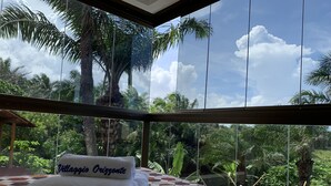 A bed with a towel labeled "Villaggio Orizzonte" and a view of lush greenery and palm trees through large windows. - Villaggio Orizzonte (Salvador)
