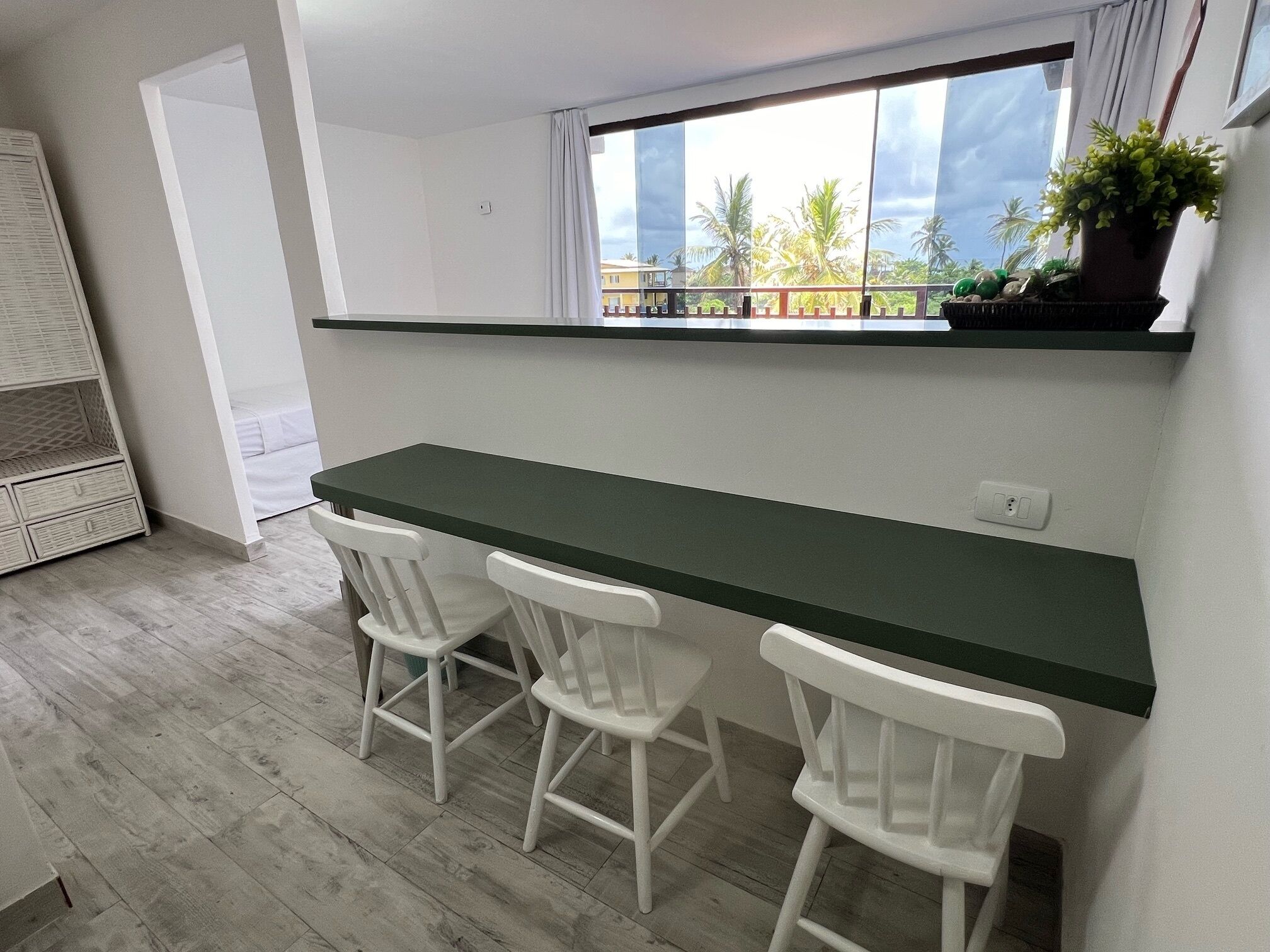 Una pequeña cocina americana con encimera verde oscuro, tres sillas blancas y ventana con vistas a un balcón con plantas y palmeras.