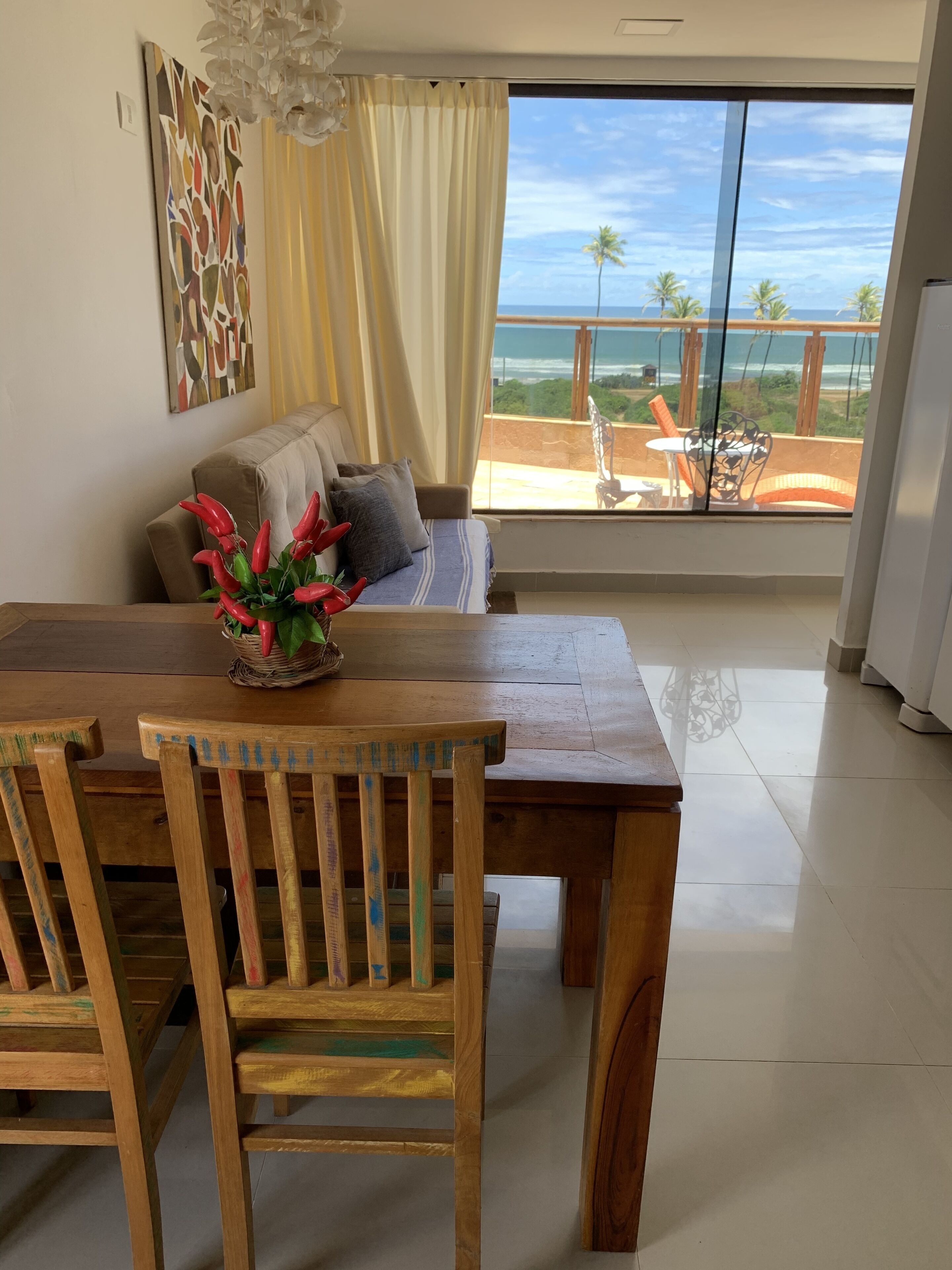 Ein Essbereich mit Holztisch und -stühlen, eine Couch mit Blick auf den Strand und ein großes Fenster mit Vorhängen.