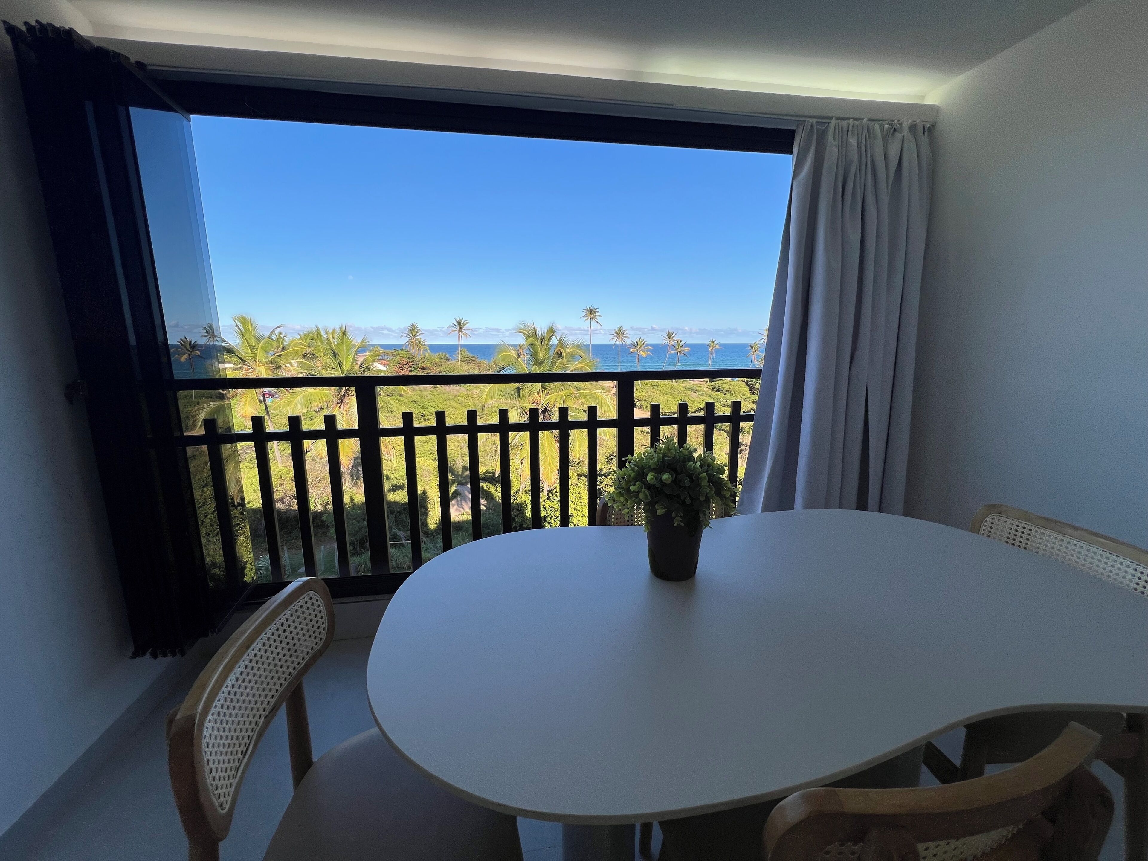 A balcony with a table and chairs, a potted plant, and a view of the ocean and palm trees.