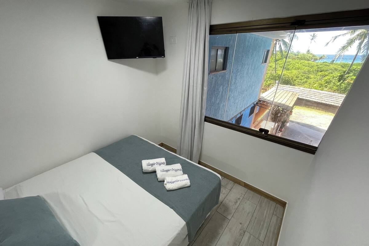 A hotel room with a bed, a flat-screen TV mounted on the wall, a window with a view of a blue building and greenery, and a small balcony.