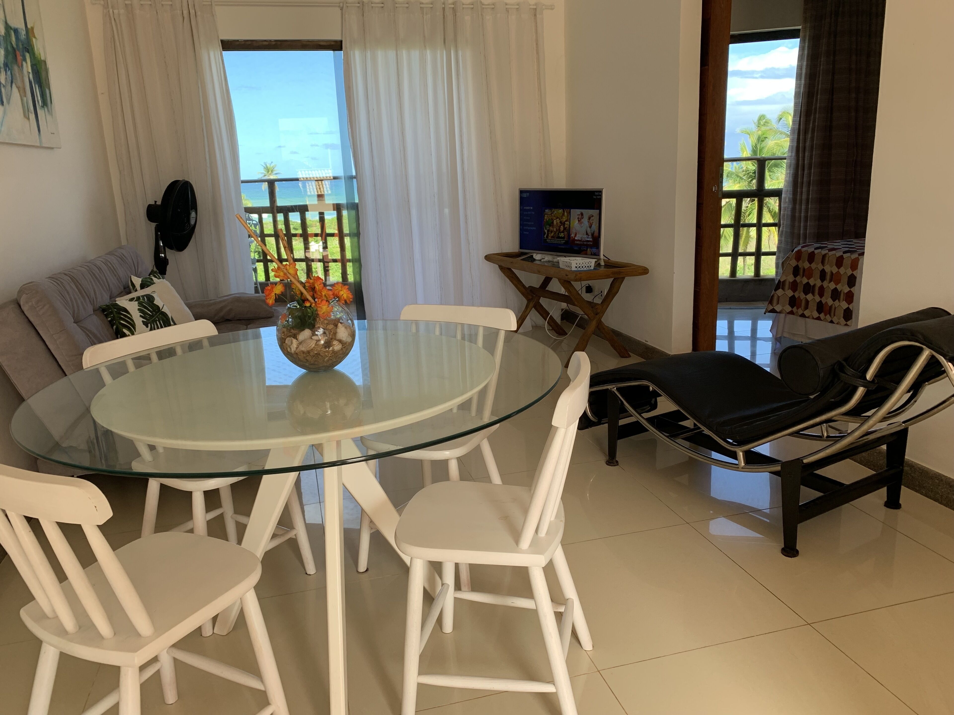 Un salon avec une table en verre, des chaises blanches, un canapé, un téléviseur et une vue sur l’extérieur.
