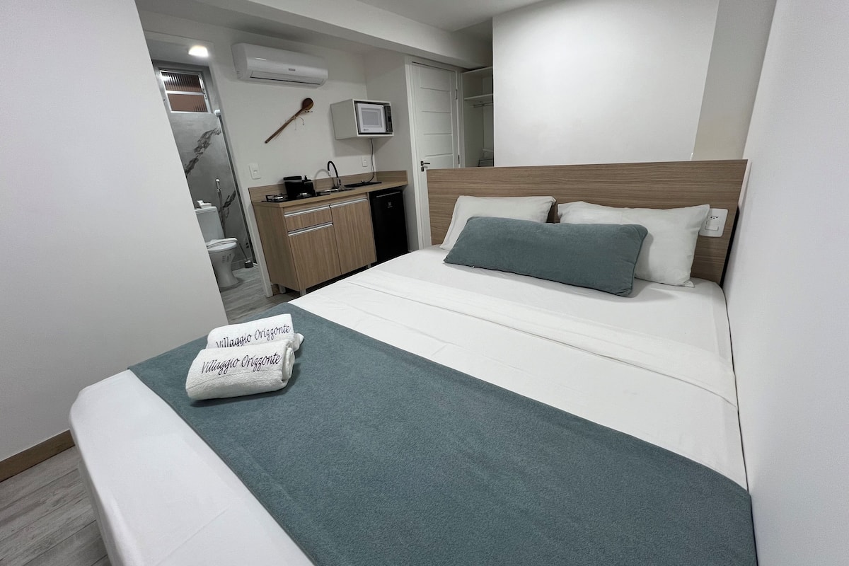 A hotel room with a bed, a bedside table, a small desk, a chair, a window, and a bathroom visible through an open door.