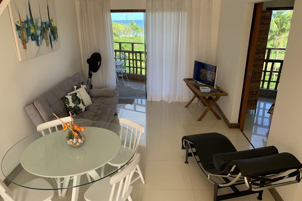 A living room with a glass table, white chairs, a grey sofa, a black bench, a TV on a wooden stand, and a view of the outdoors.