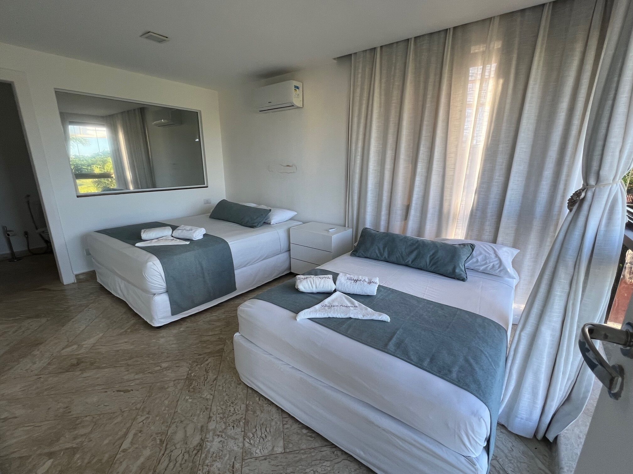 A hotel room with two beds, a large mirror, a window with curtains, and an air conditioning unit.