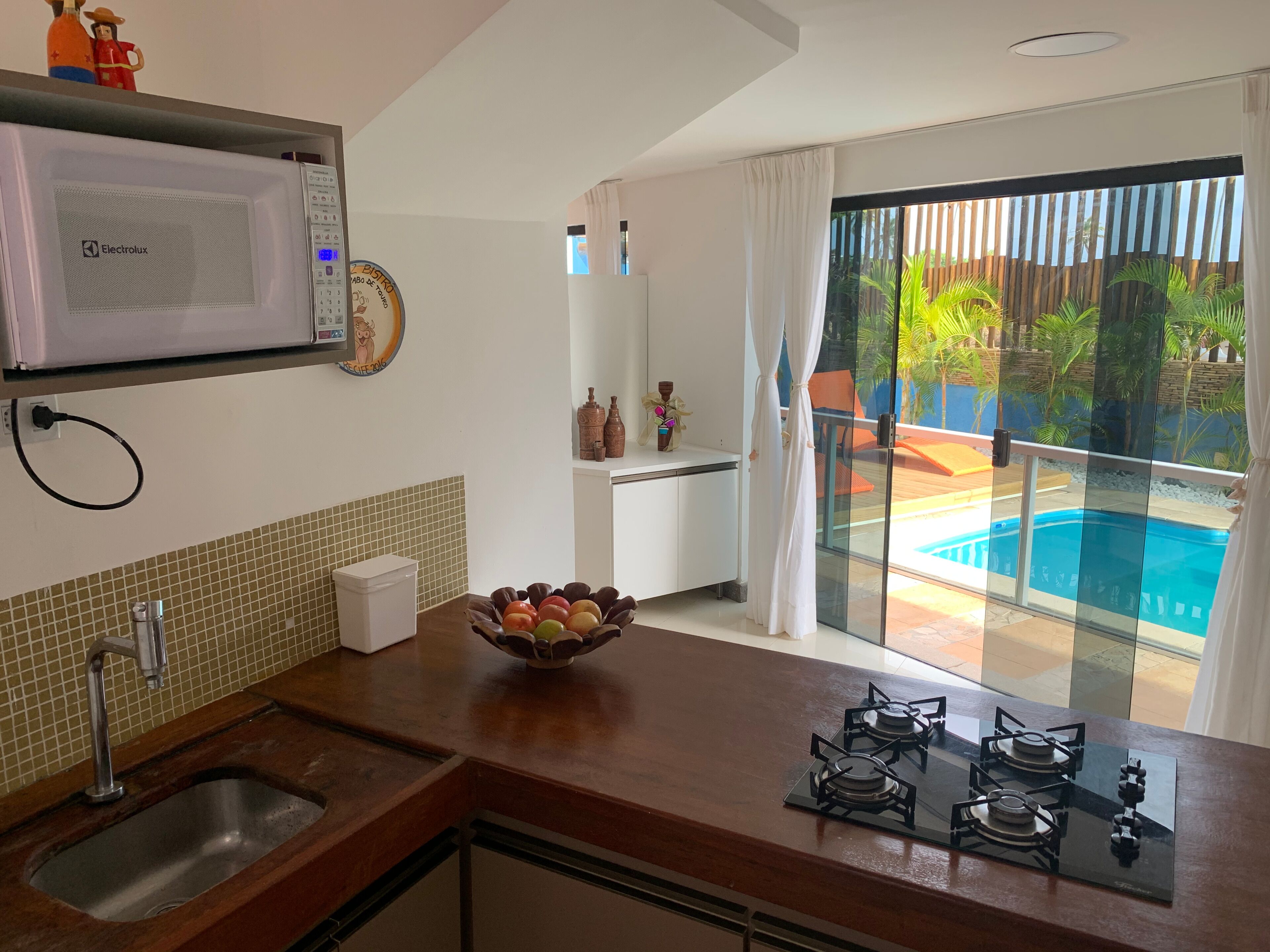 A modern kitchen with a microwave, sink, and gas stove. A bowl of fruit is on the counter, and there's a view of a pool through a glass door.