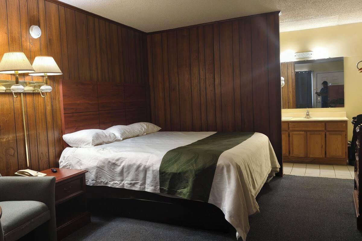 A hotel room with a wooden paneled wall, a bed with white bedding, a desk with a lamp, a chair, and a bathroom with a mirror and sink.