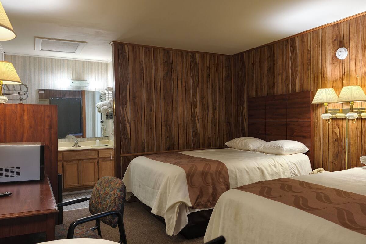 A hotel room with two beds, a desk with a lamp, a chair, and a bathroom visible in the background.