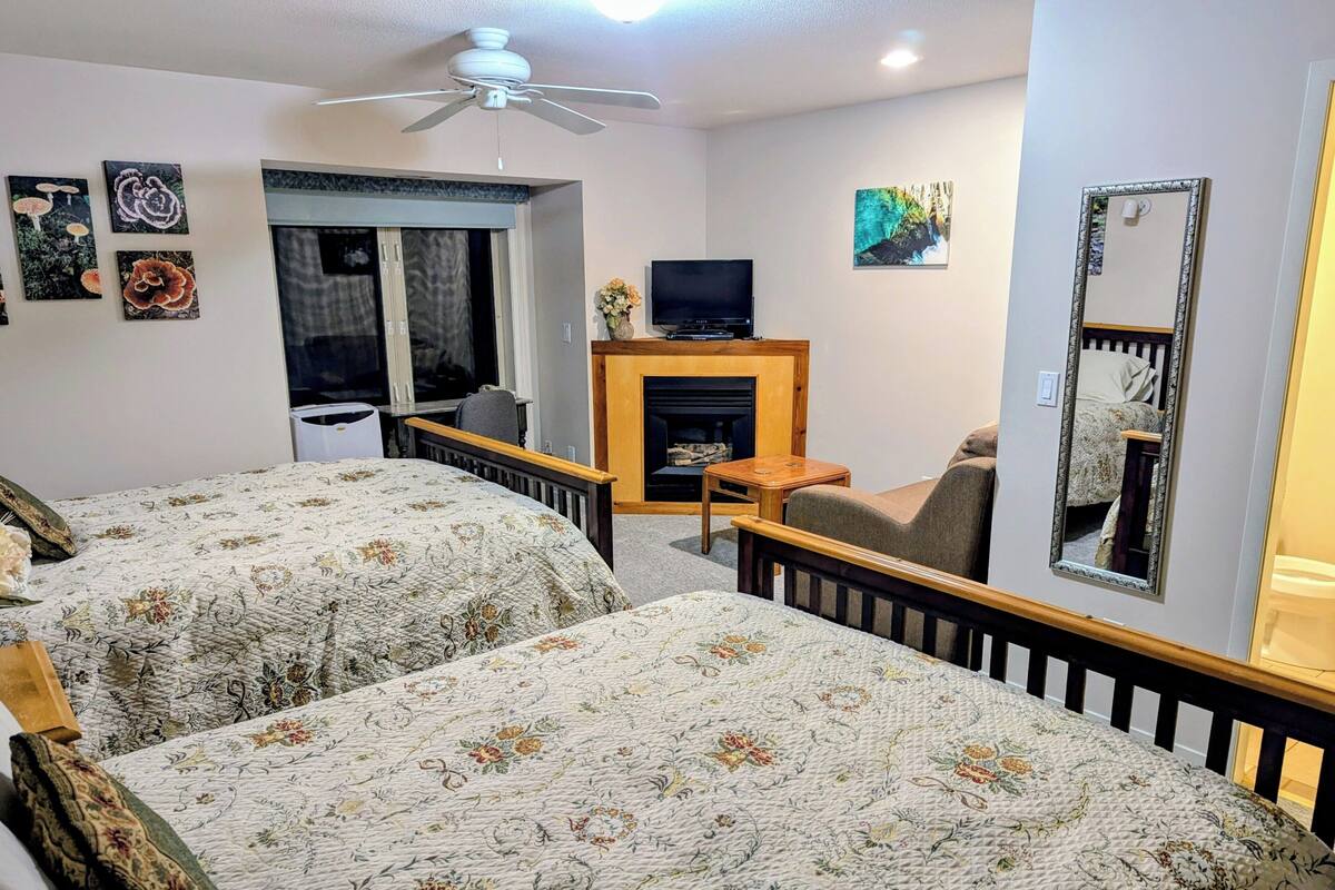 A hotel room with two beds, a ceiling fan, a TV, a fireplace, and artwork on the walls.