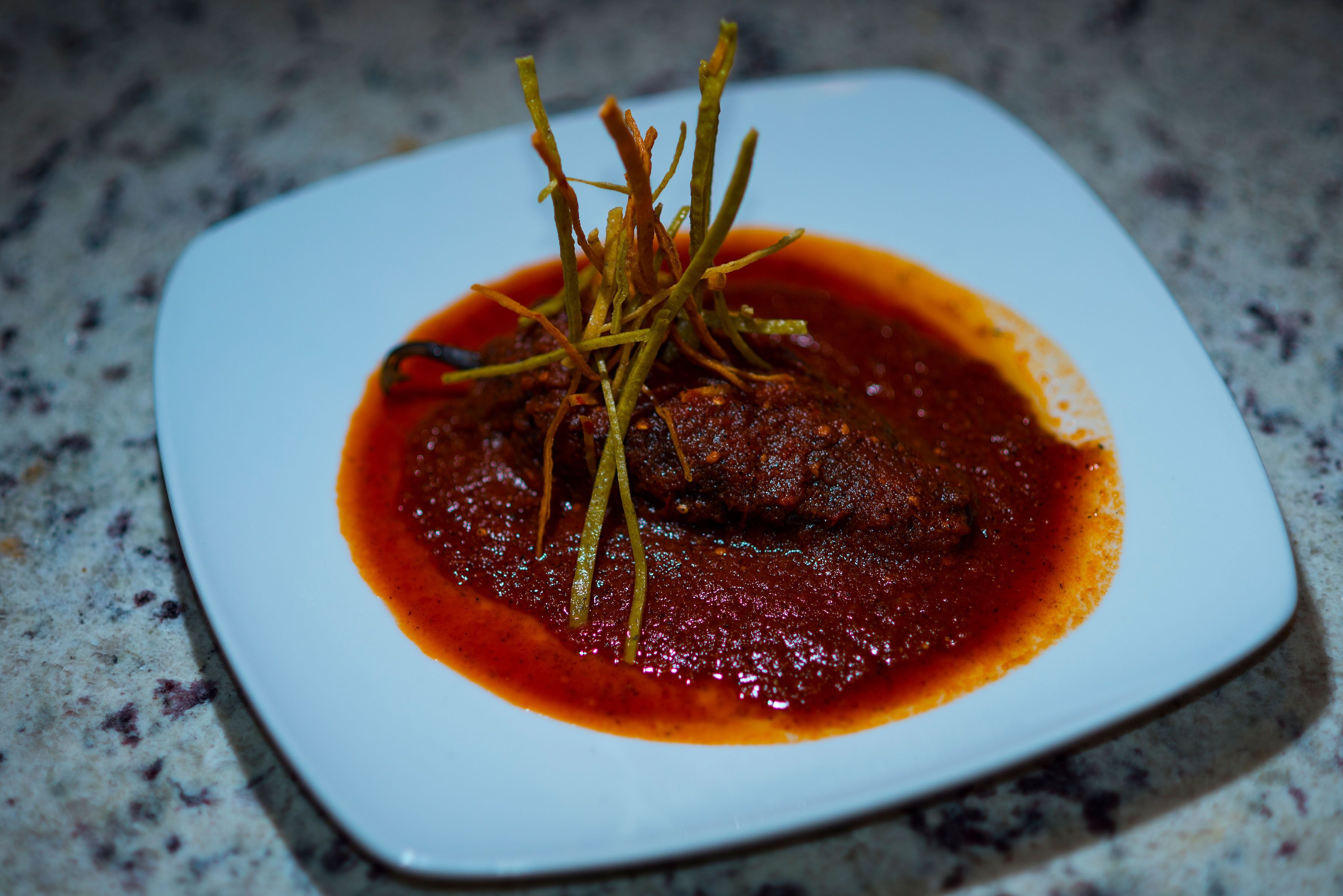 A square plate with a meat dish in a red sauce, garnished with thin strips, on a speckled countertop.