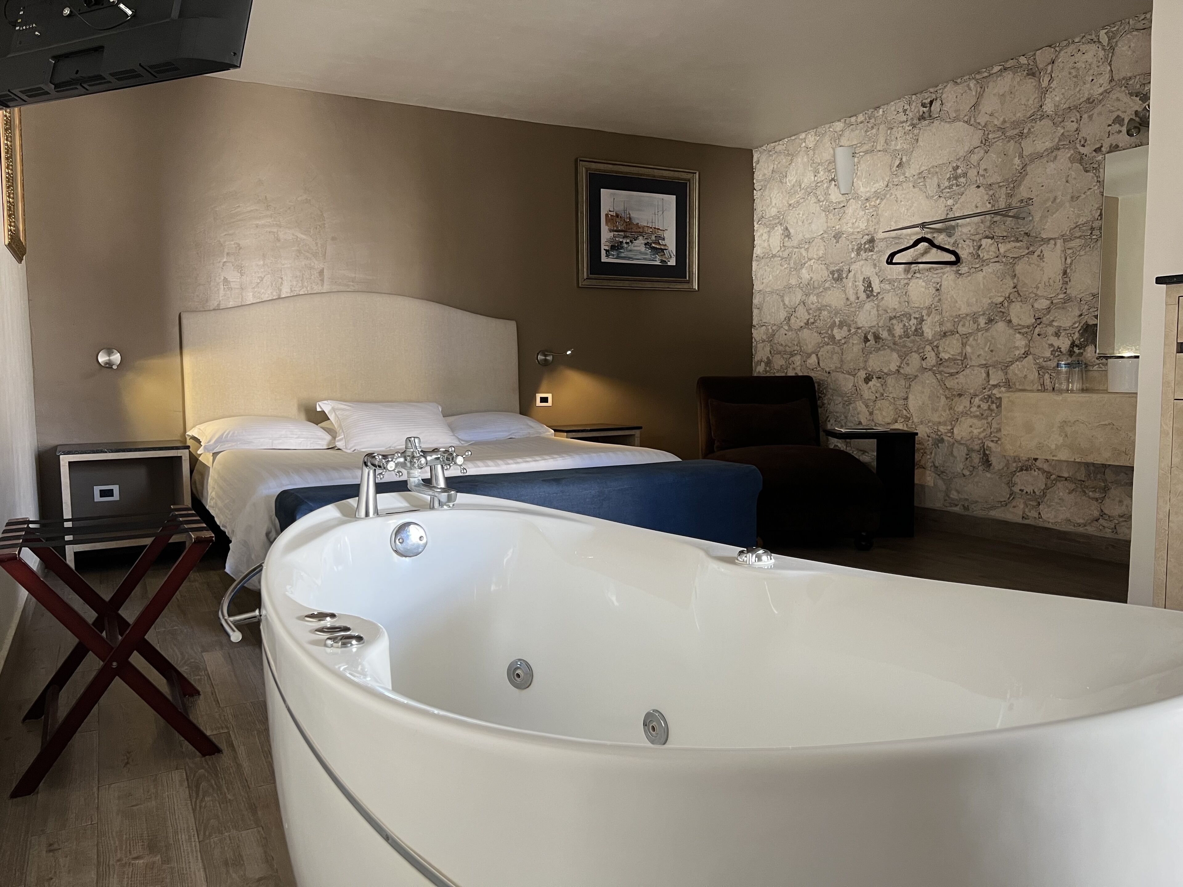 A hotel room with a large bathtub, two beds, a stone wall, a framed picture, and a small table with a lamp.