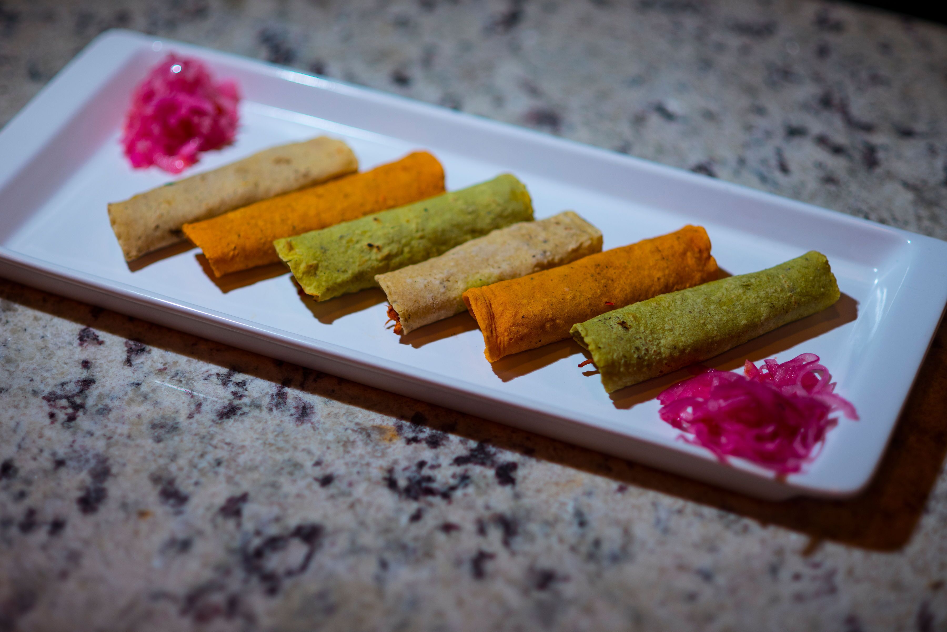 A white rectangular plate with six rolled items in various colors, placed on a granite countertop.