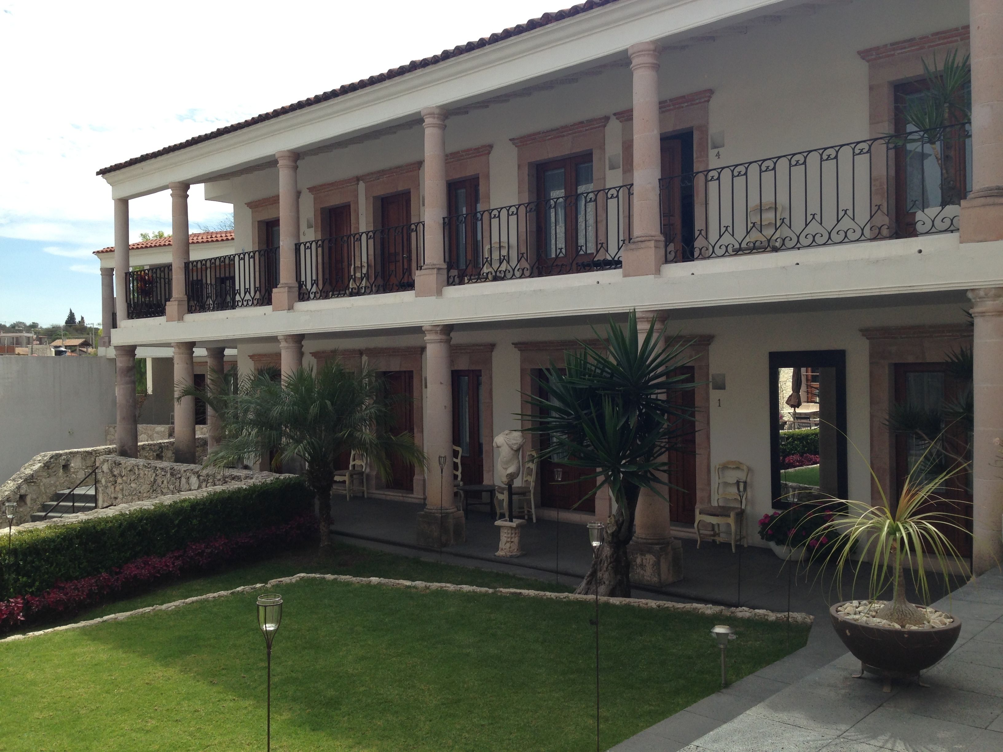 Un edificio de dos pisos con un balcón, columnas y un jardín bien cuidado.