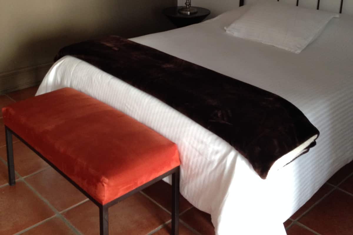 A bed with a white mattress and a dark blanket, a round mirror on the wall, a bedside lamp, and a red bench.