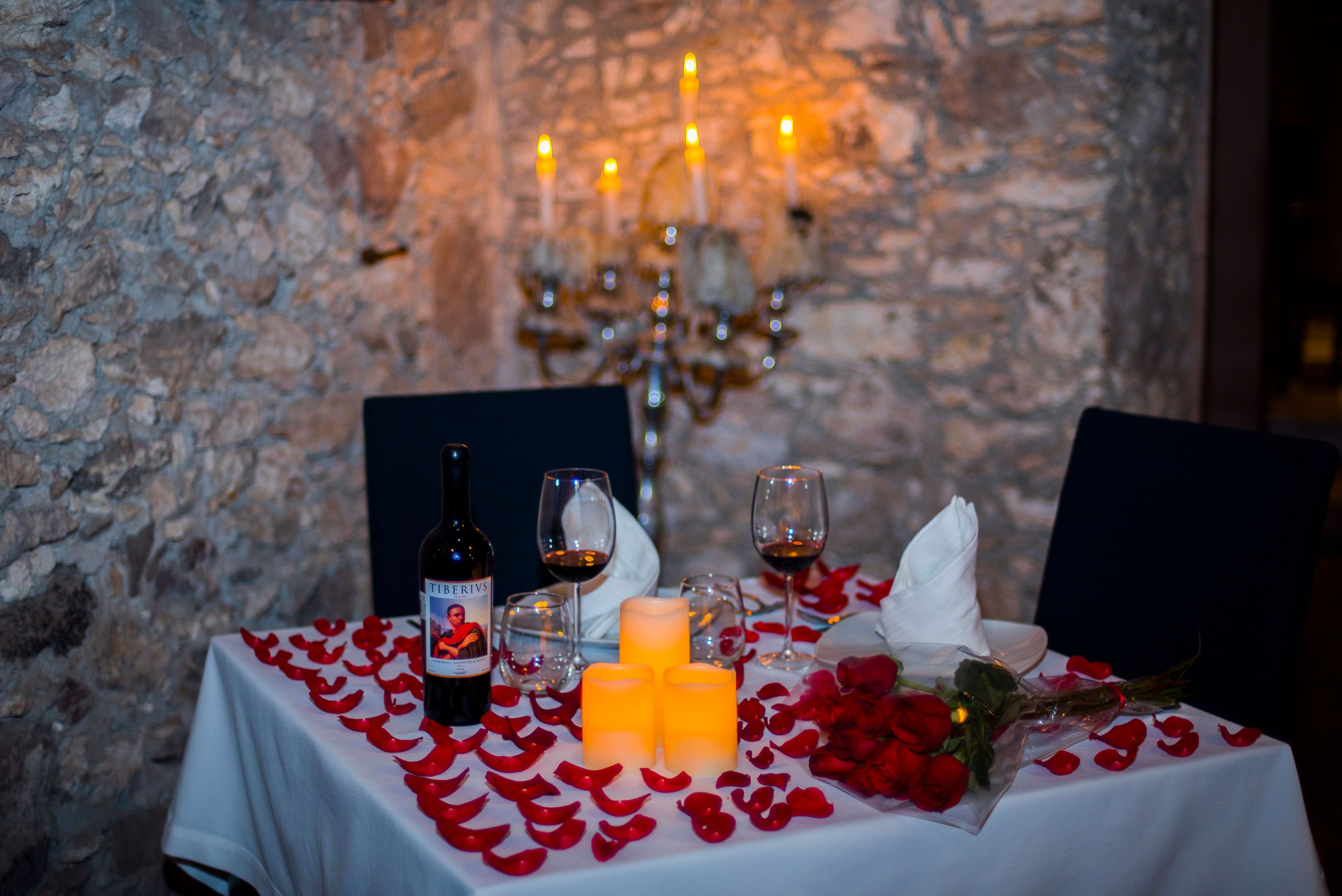 Una mesa dispuesta con velas, copas de vino, una botella de vino y pétalos de rosa.