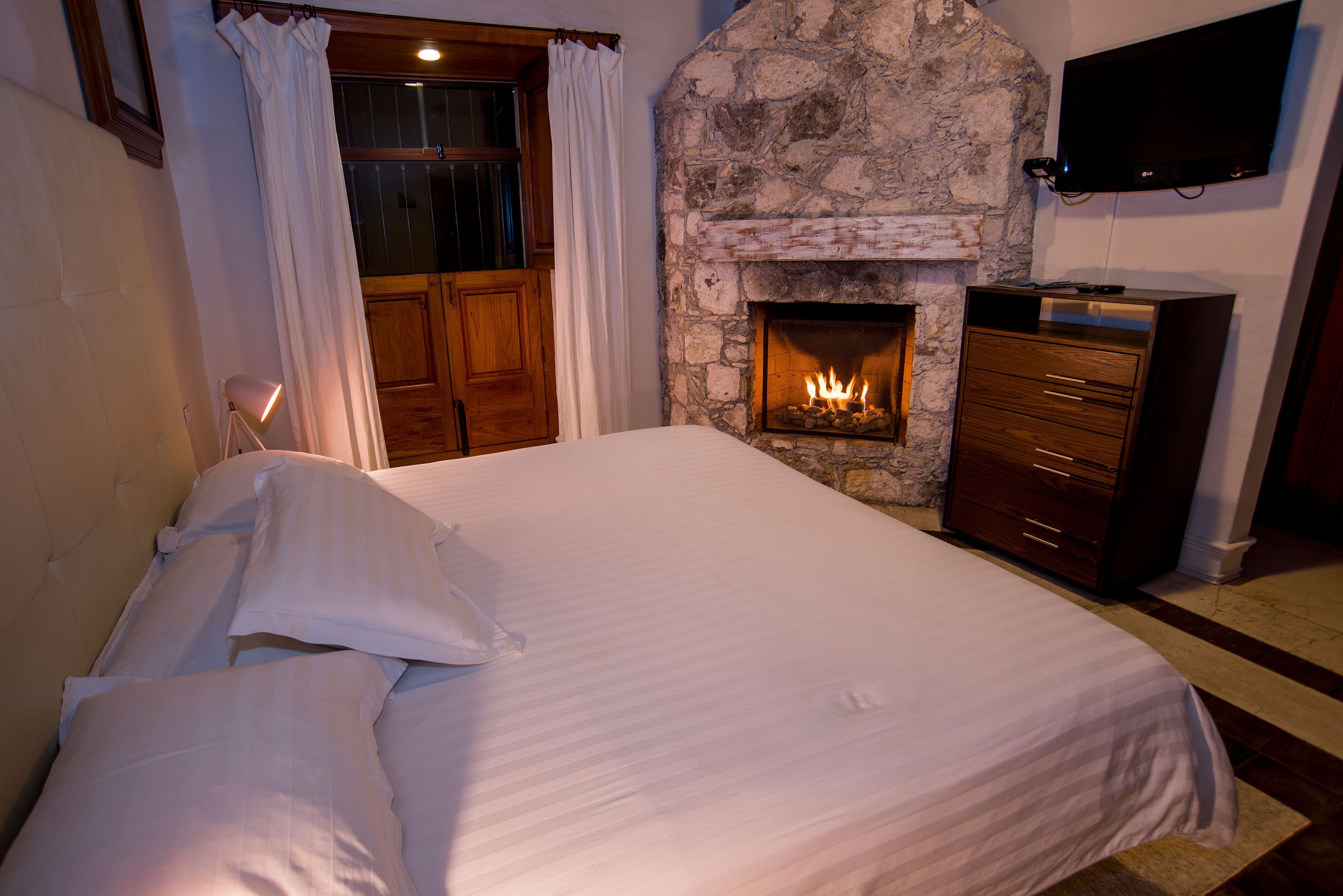 A bedroom with a stone fireplace, a bed with white linens, wooden furniture, and a television mounted on the wall.