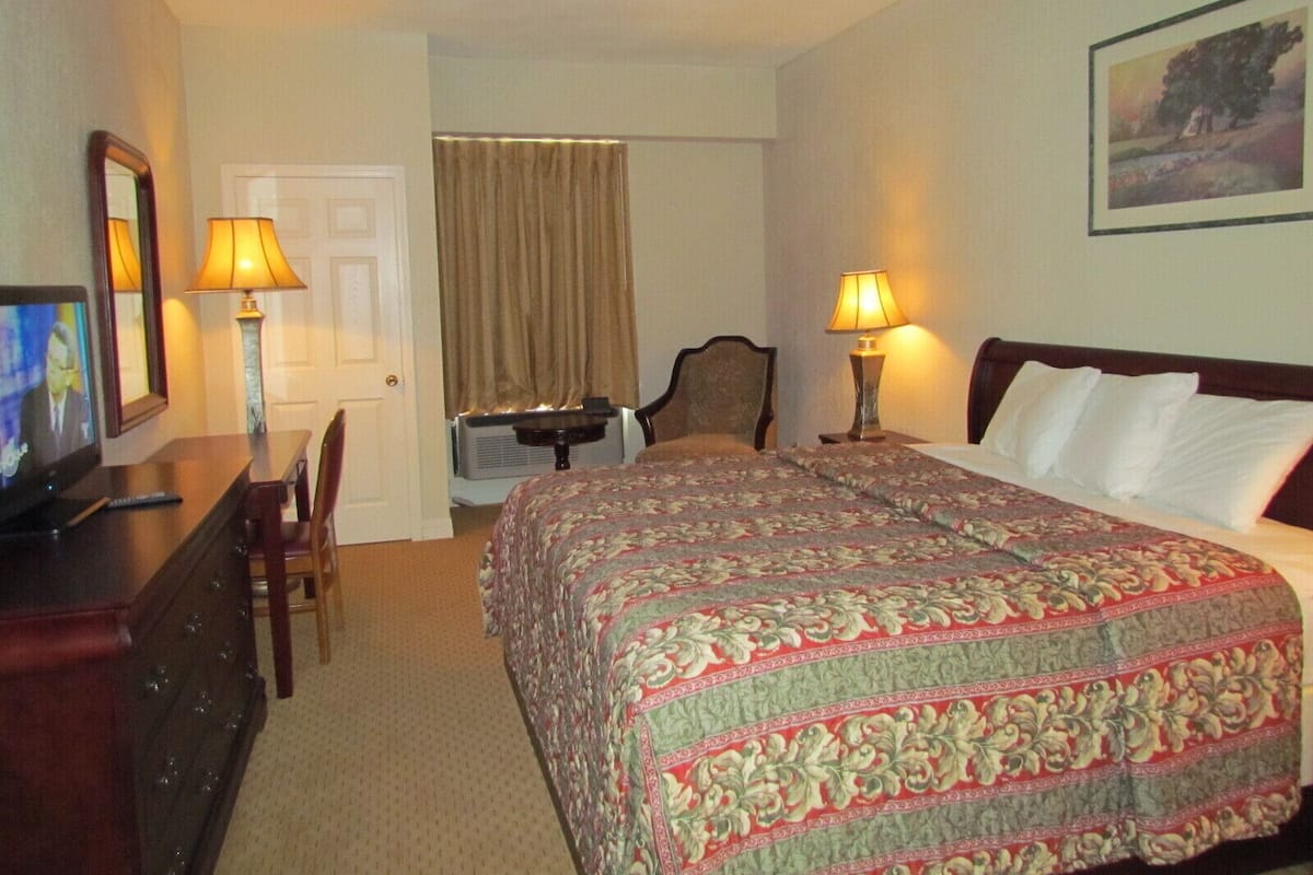 A hotel room with a bed, a desk with a lamp, a chair, a television, and a painting on the wall.