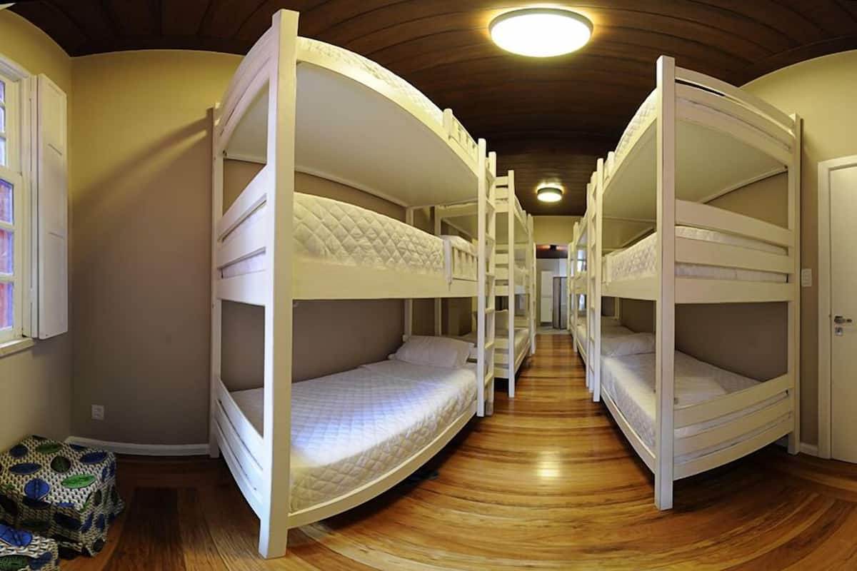 Quarto com beliches dispostos em um corredor, pisos de madeira e uma luminária no teto.