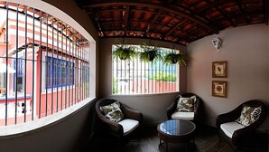 Una habitación de esquina con sillones de mimbre, una mesita redonda, una ventana con plantas y vistas a un edificio rojo.