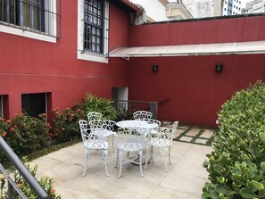 Un patio con sillas y mesa de metal blanco, rodeado de muros rojos y follaje.