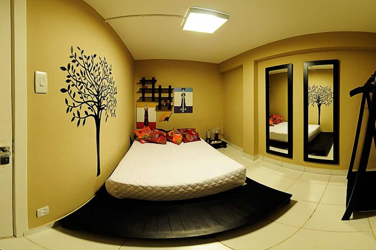 Quarto com cama, decalque de árvore na parede, espelho e prateleira com objetos decorativos.