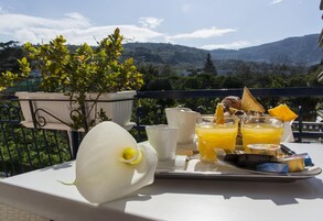 Ein Frühstückswagen mit Orangensaft, Toast und einer weißen Blume auf einem Balkon mit Bergblick.