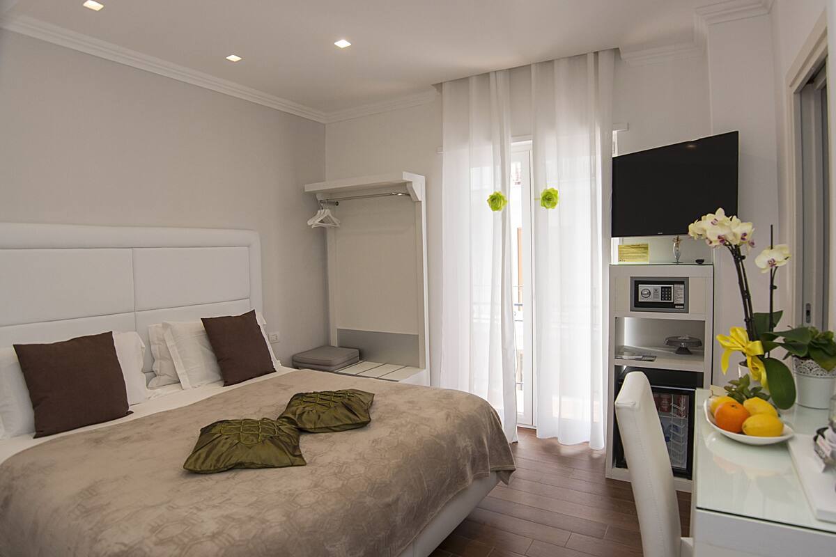 A modern hotel room with a large bed, a desk with a computer, a TV, and a small kitchen area.